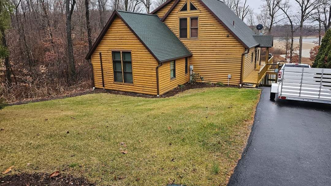 Yellow log cabin with green roof, set in a yard, next to a black asphalt driveway, and a white trailer.