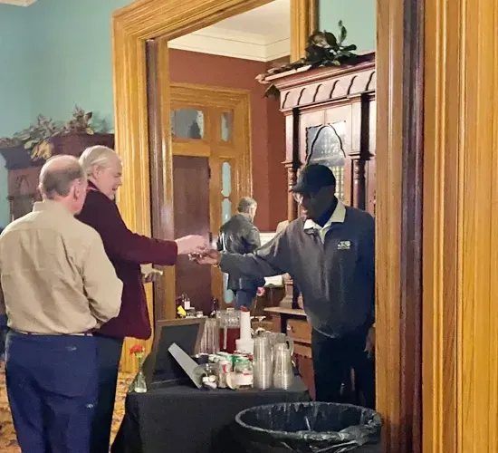 Two people shake hands across a tray table in a doorway, with a fireplace in the background.