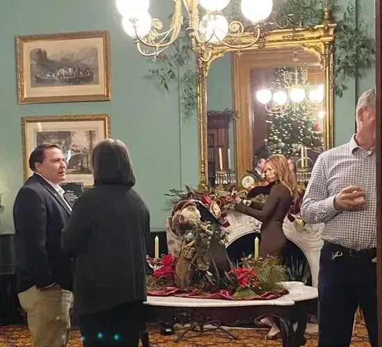 People mingling in an ornate room with a chandelier, large mirror, and floral centerpiece.