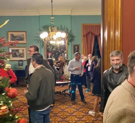 People mingling in a warmly lit room with a chandelier, patterned carpet, and holiday decorations.