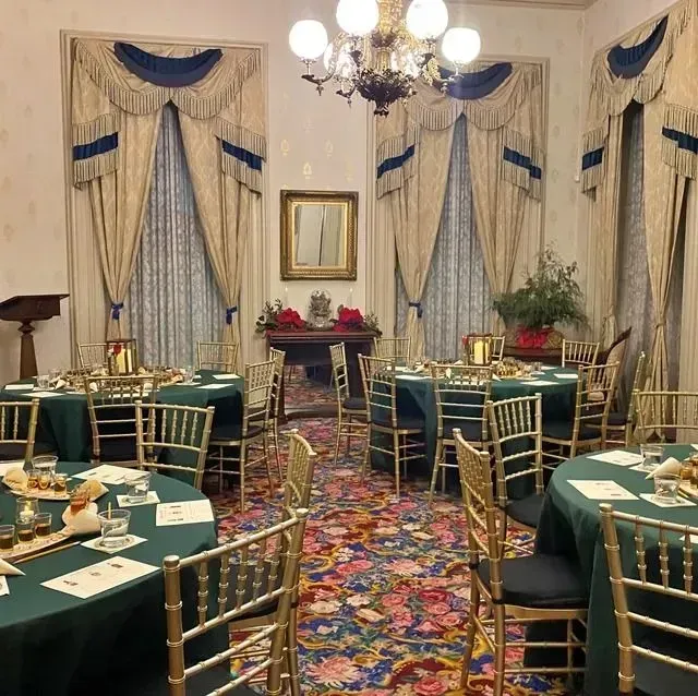 Elegant banquet room with green-clothed tables, gold chairs, patterned carpet, and tall draped windows