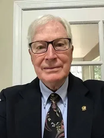 Man in a dark suit and patterned tie standing indoors, wearing glasses and smiling softly.