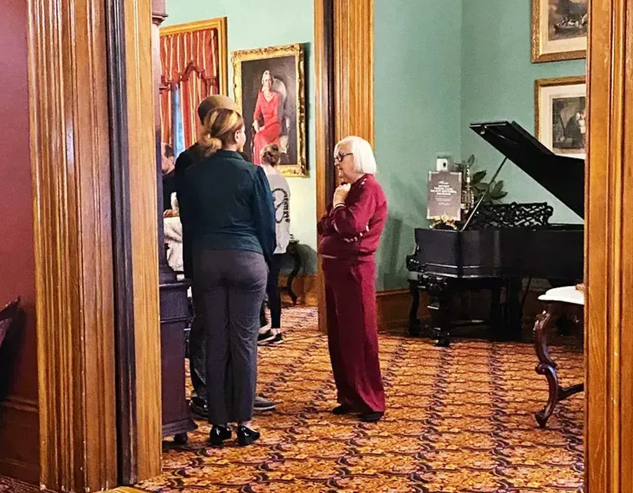 Two people converse in a gallery room with framed portraits, green walls, and a grand piano.
