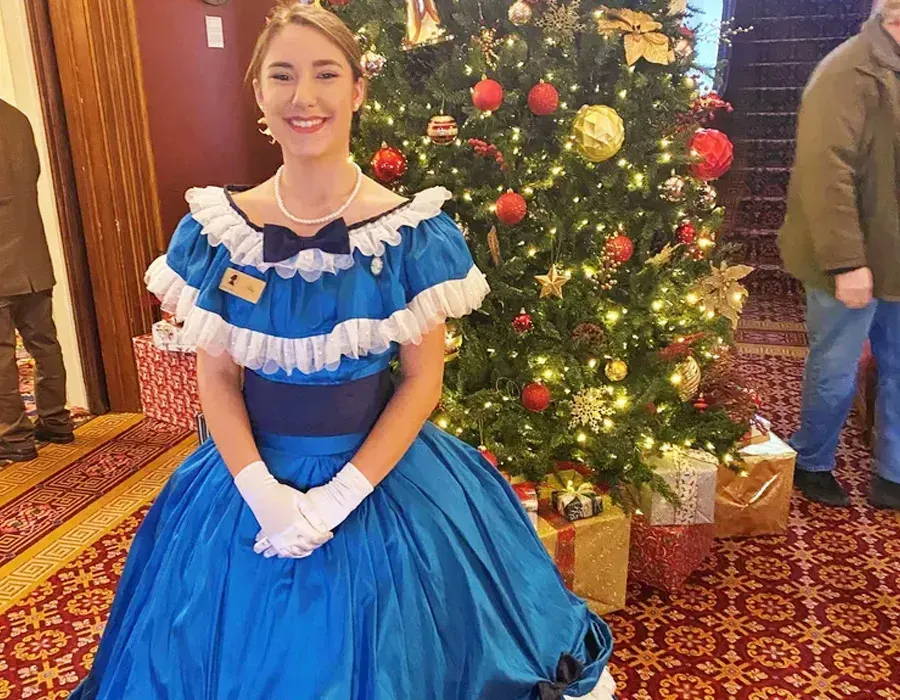 Woman in a blue dress posing beside a decorated Christmas tree indoors.