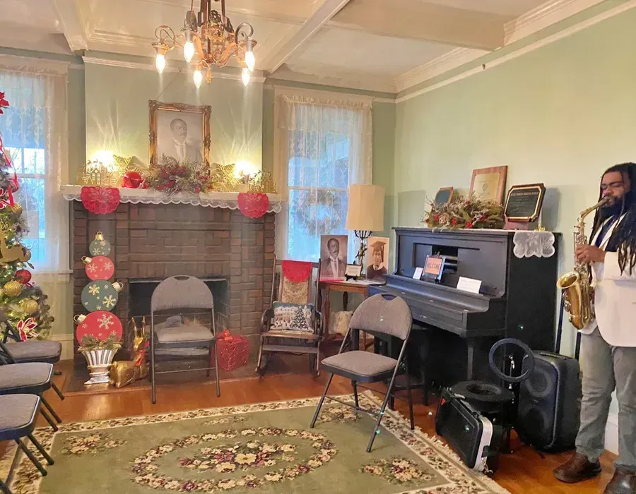 Cozy living room with fireplace, holiday decor, piano, chairs, and a man playing saxophone near the wall.