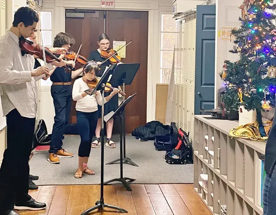 Children playing violins in a hallway near a decorated Christmas tree and music stands.
