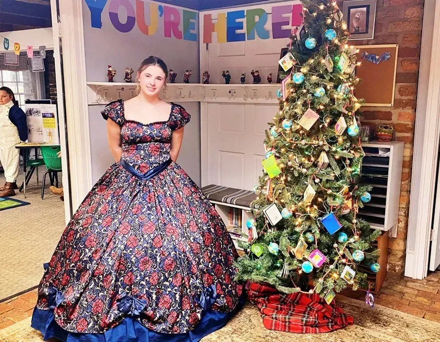 Woman in a colorful floral gown beside a decorated Christmas tree and fireplace in a festive room