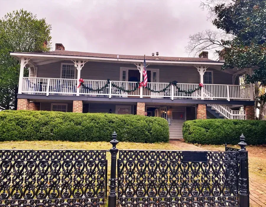 Historic two-story house with white porch, American flags, hedges, and ornate black fence
