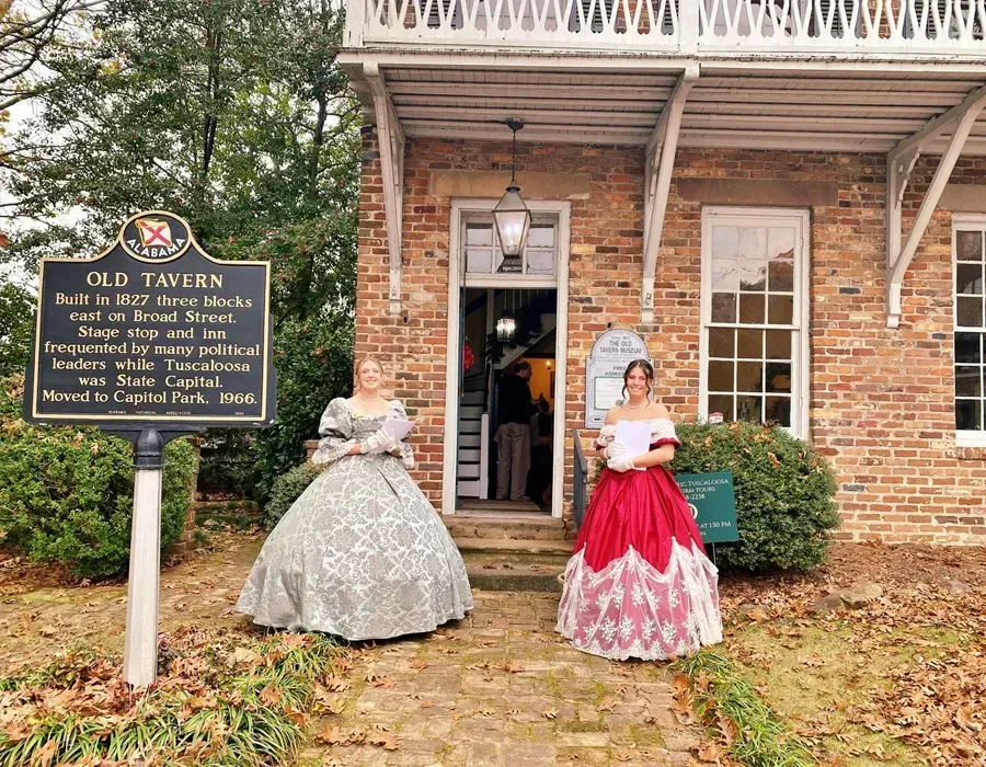 Two women in historic dresses outside Old Tavern, one holding a paper, brick building and sign nearby