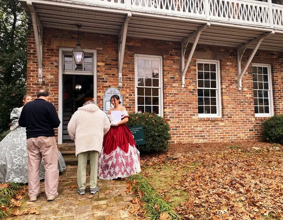 Three people stand outside a brick building; one wears a red dress and bonnet, holding papers.
