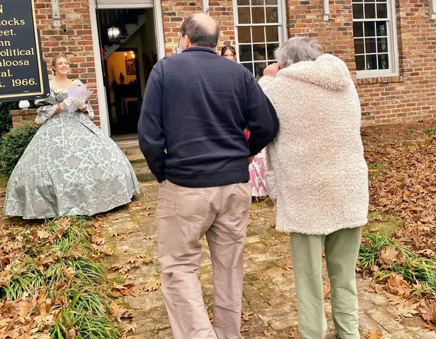 People standing outside a brick building on a leaf-covered path, one in a gray dress near the doorway.