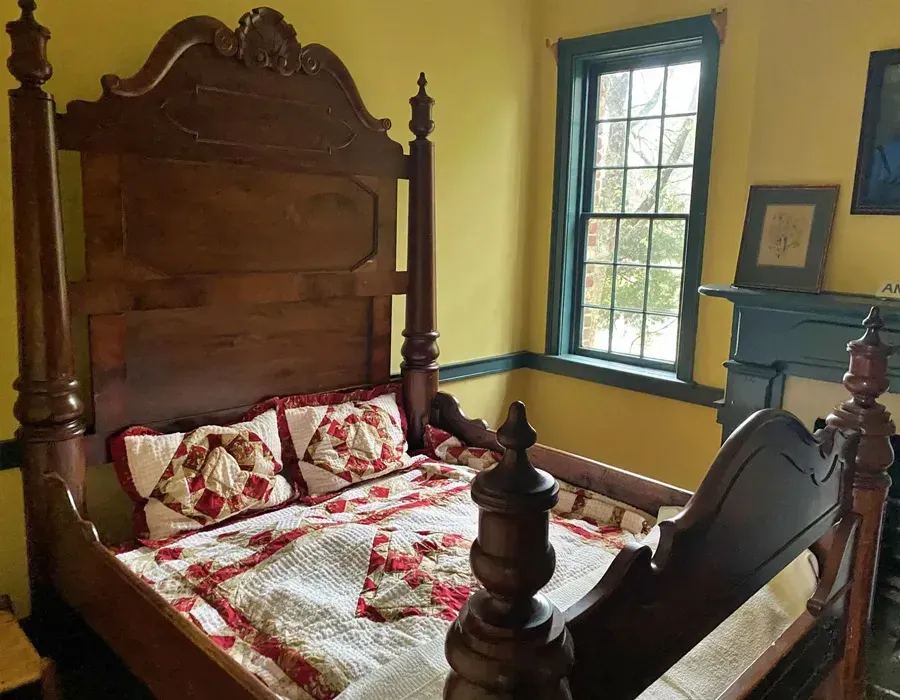 A historic four-poster bed with red quilt and pillows in a yellow bedroom with blue-trimmed windows.