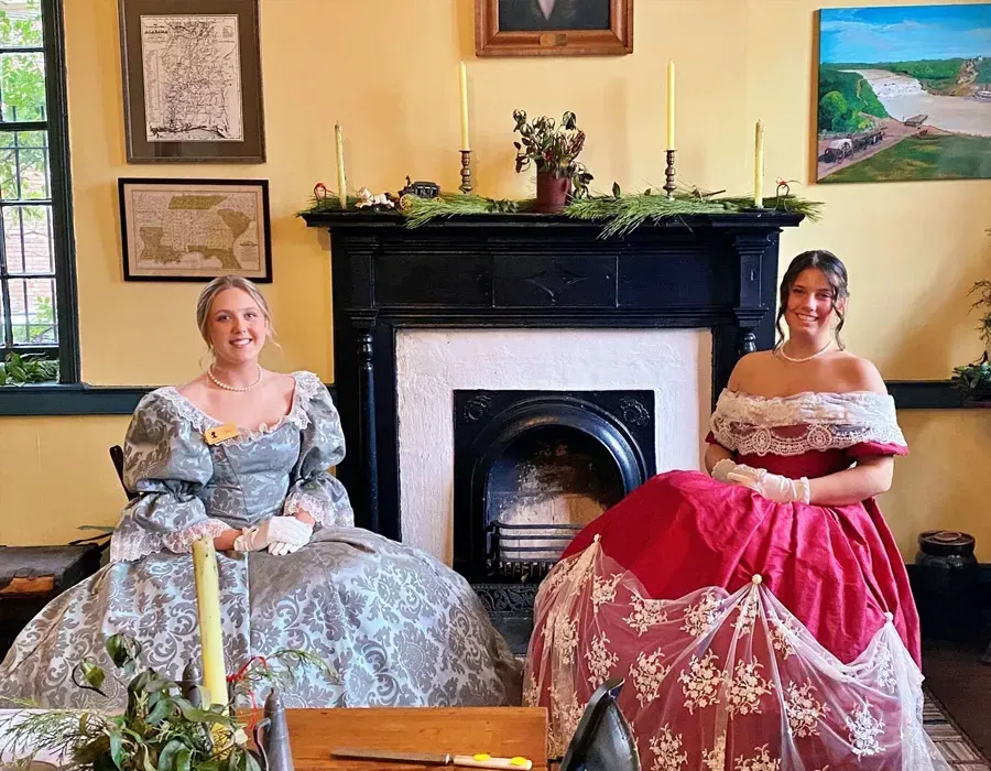 Two women in ornate ball gowns seated in a parlor by a fireplace