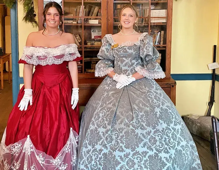 Two women in ornate red and gray historical gowns standing indoors in front of a wooden cabinet.