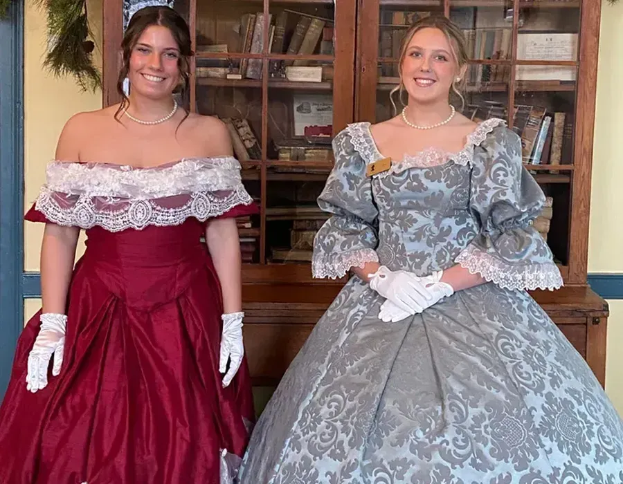 Two women in formal gowns and white gloves pose indoors in front of a wooden cabinet.