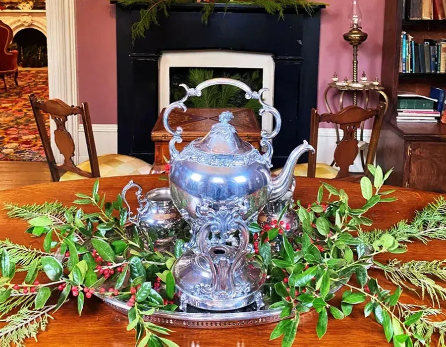 Silver teapot centerpiece on a wooden table, framed by green branches in a cozy room.