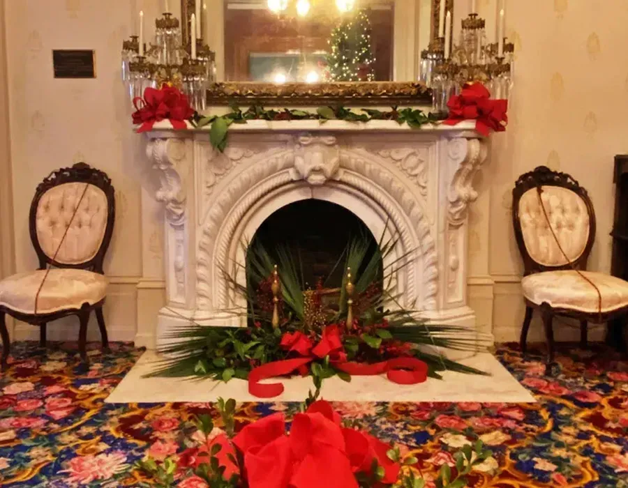 Ornate white fireplace with holiday decorations, red bows, and two chairs in a colorful room