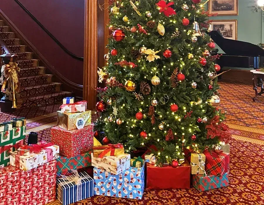 Decorated Christmas tree beside a staircase, with wrapped gifts on a patterned carpet in a cozy room