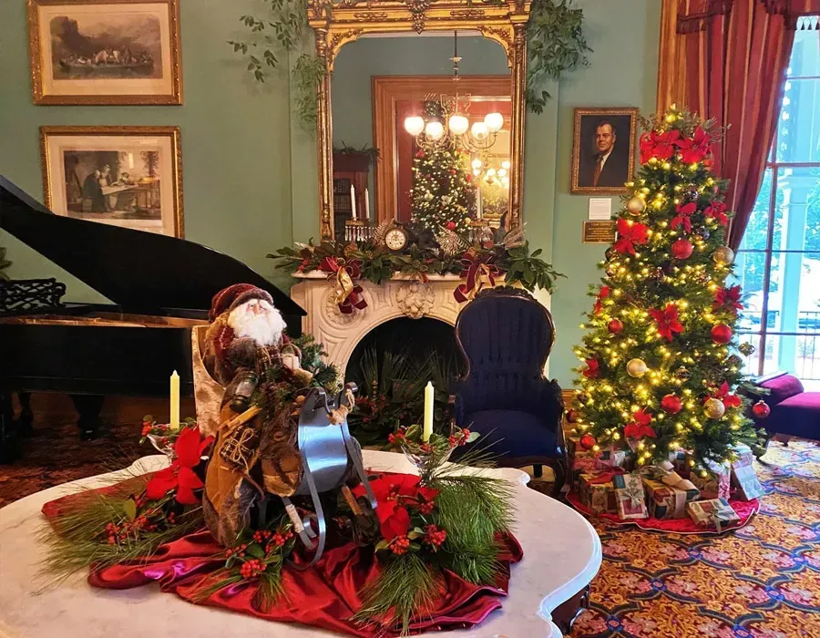Festive living room with Christmas tree, piano, fireplace, and Santa figures on a decorated table.