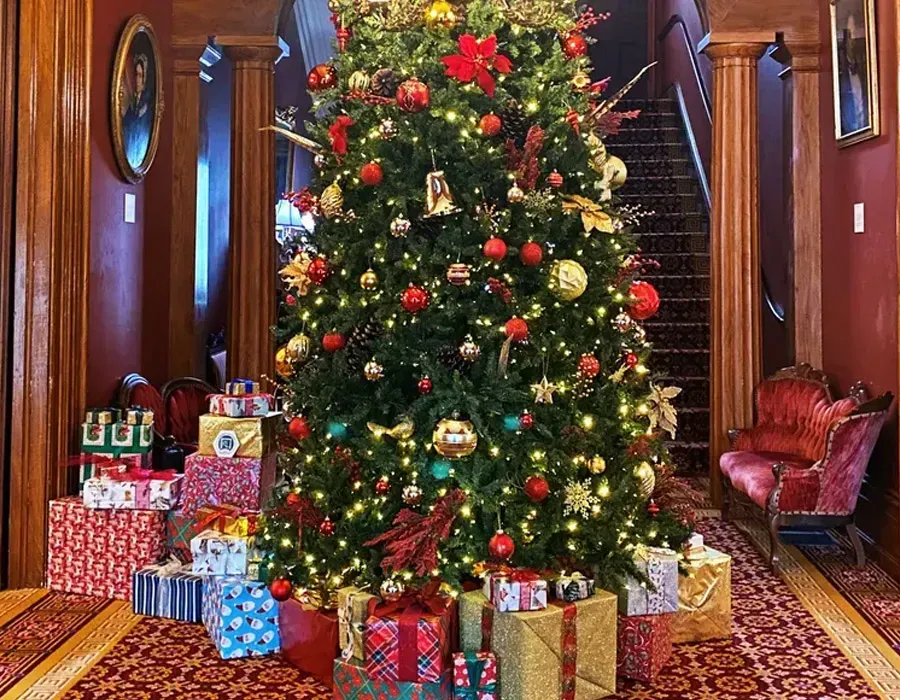 Christmas tree in a grand hallway, decorated with lights and ornaments, surrounded by wrapped gifts.