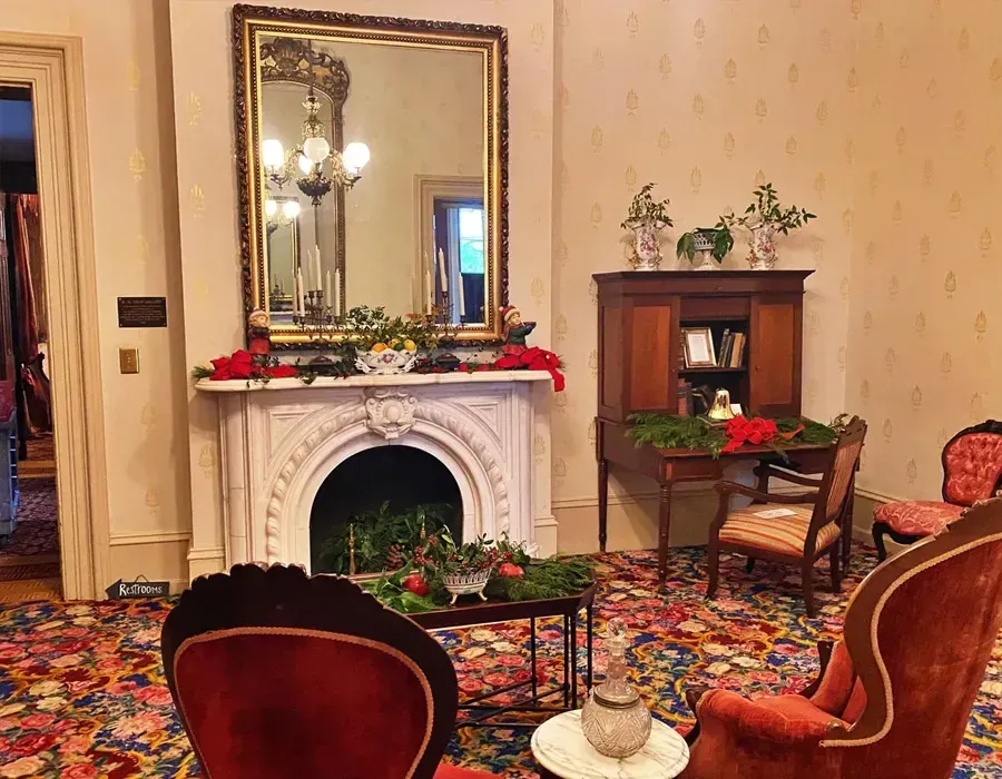 Elegant parlor with ornate white fireplace, mirror, red chairs, and floral decor on patterned carpet.
