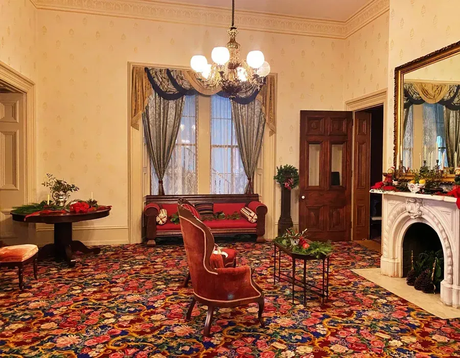 Elegant vintage sitting room with floral carpet, red chairs, chandelier, fireplace, and window drapes.