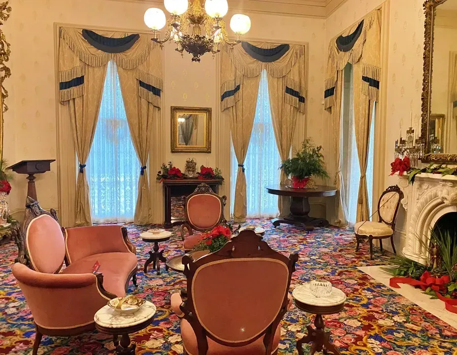 Elegant living room with pink chairs, floral carpet, tall drapes, and a fireplace with red flowers.