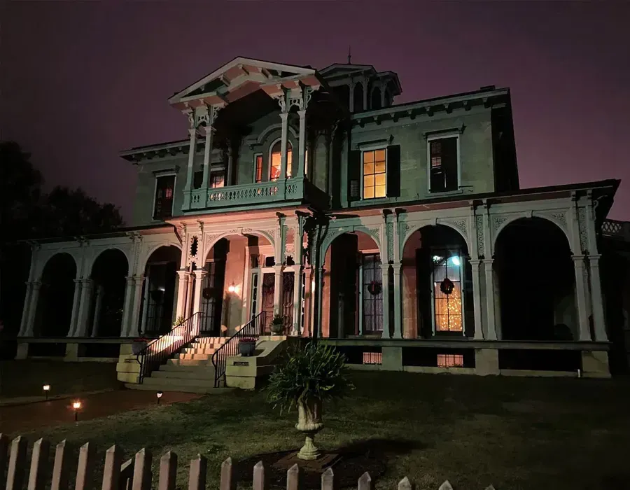Night-lit old mansion with white columns and a porch, glowing windows, and a dark purple sky.