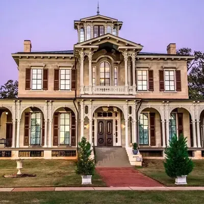 A two-story historic mansion with a wrap-around porch, ornate woodwork, and a cupola against a soft purple twilight sky.