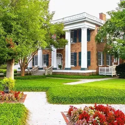 A historic two-story brick mansion with white columns, set behind a manicured lawn and garden beds with red flowers.