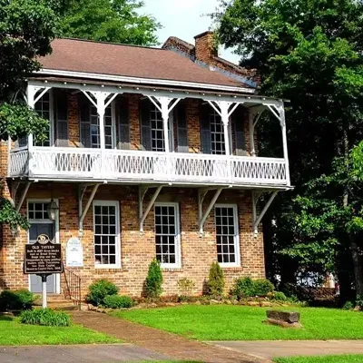 A historic, two-story stone house with a white wooden balcony, nestled among trees on a grassy lot.