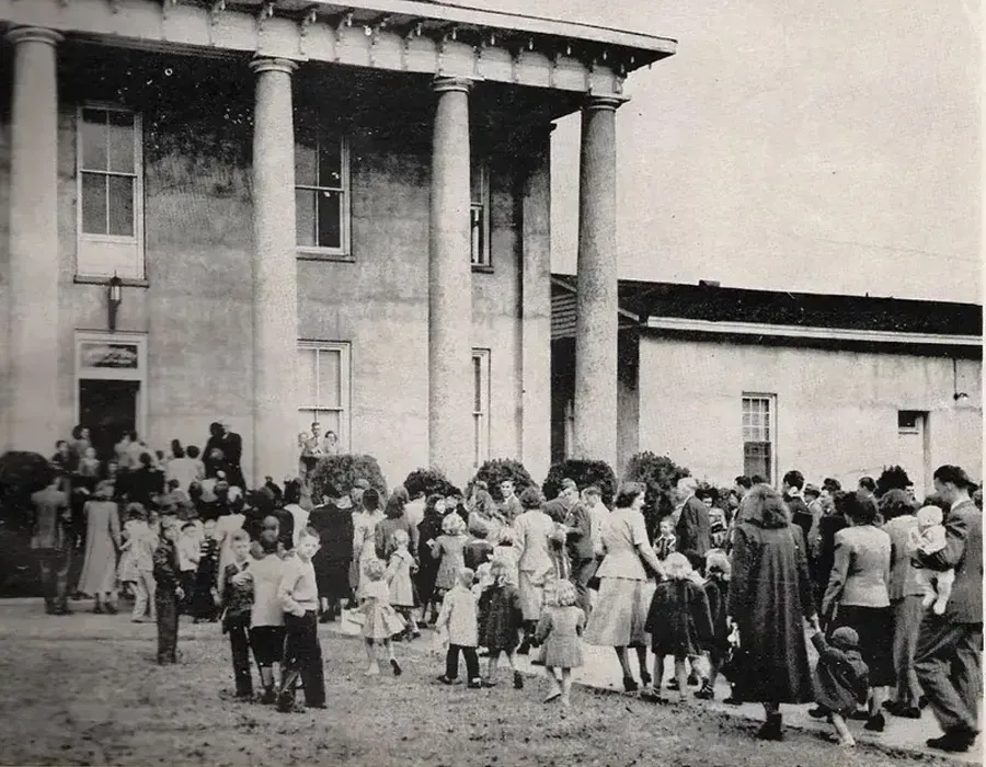 Crowd gathered outside a columned building, standing in line in a dusty courtyard.