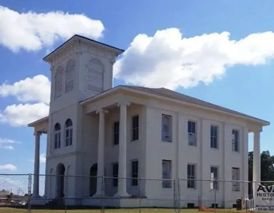 White two-story building with a tower and columns, seen behind a fence under a blue sky