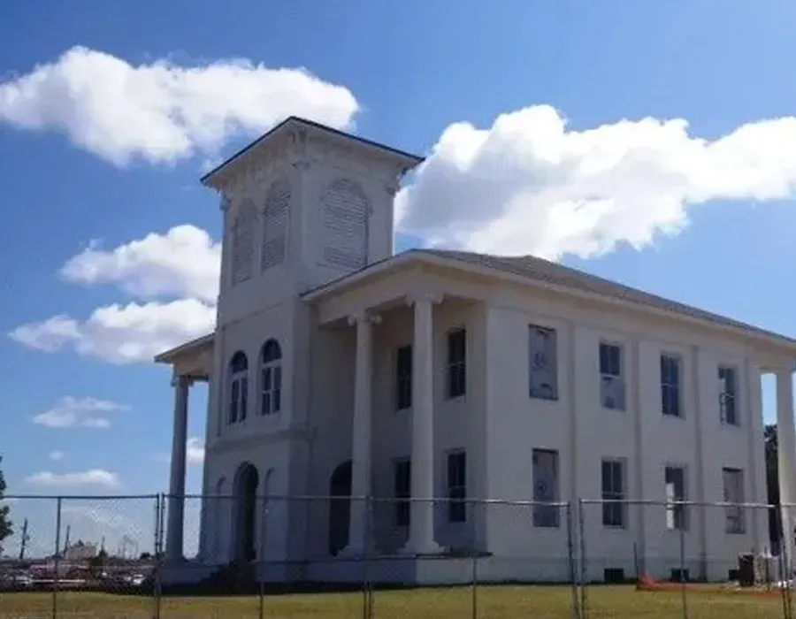 White columned building with a tall tower behind a fenced grassy area under a blue sky with clouds