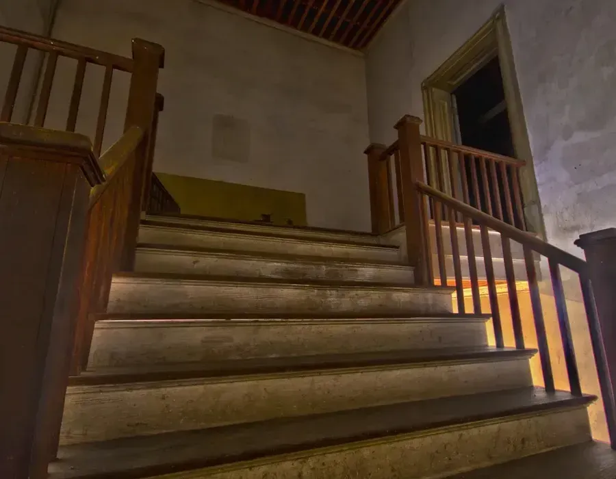 Wooden staircase in a dimly lit interior with railings and an open doorway at the top.