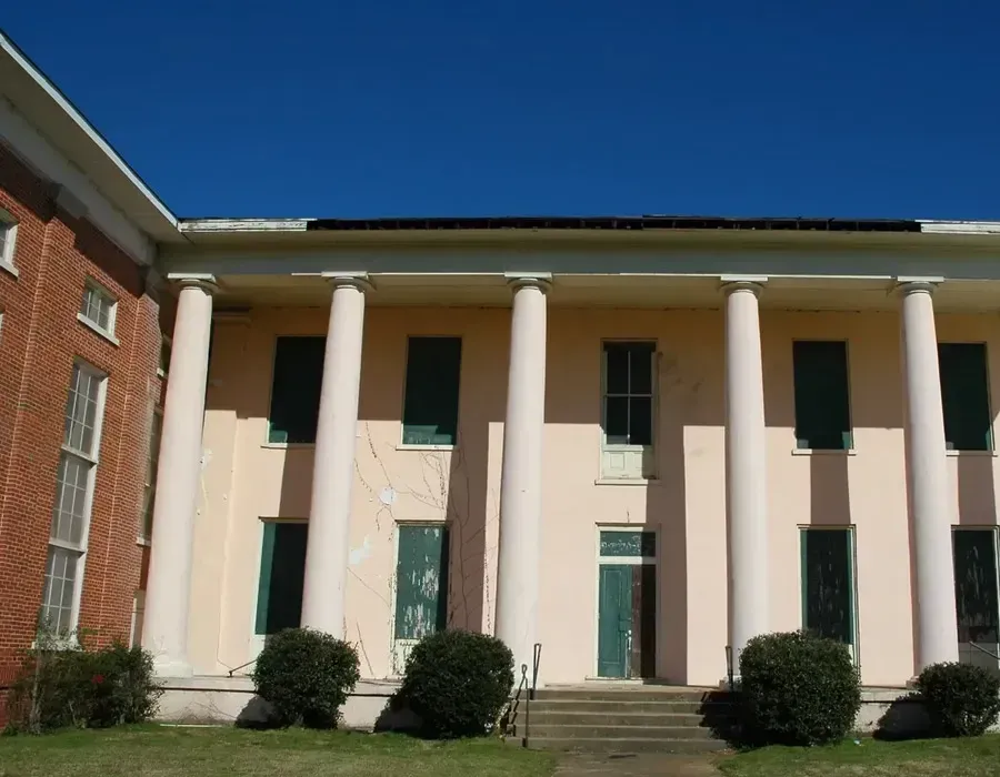 White-columned building with green shutters and a red brick wing under a clear blue sky