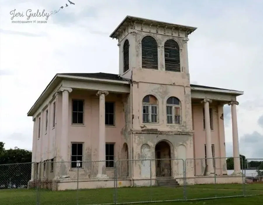 Old, pink two-story house with a tall central tower and columns, behind a chain-link fence on a grassy lot