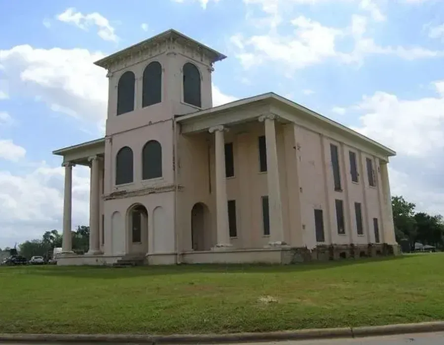 Large beige historic building with tall arched windows and a tower, seen across a grassy lawn under a blue sky