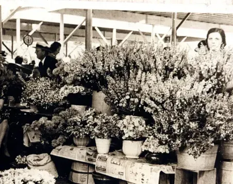 A market stall features vendors standing behind tables displaying large arrangements of flowers in pots and buckets.