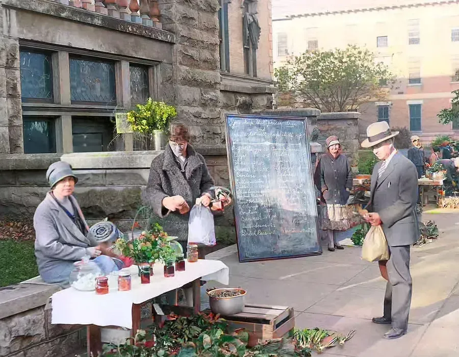 People at a sidewalk market with potted plants and a large sign in front of a stone building.