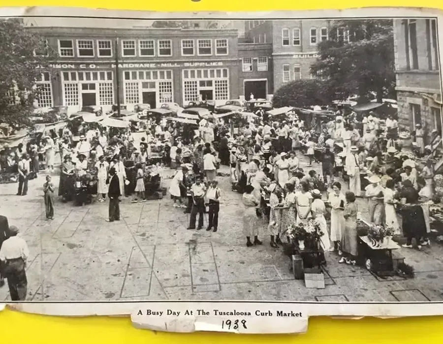 A black-and-white 1929 photo of a busy open-air street market with people gathered around stalls in front of brick buildings.