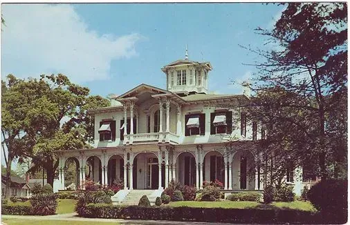 Victorian-style mansion with white columns and wraparound porch amid lush trees and lawn