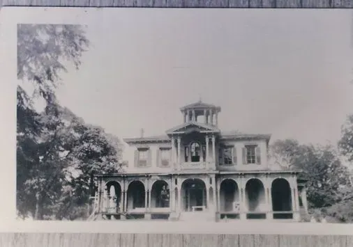 Black-and-white photo of a large two-story historic mansion with arched porches and a central tower.