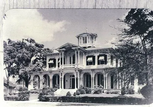 Historic Victorian mansion with wraparound porch and tower, surrounded by trees in black and white.