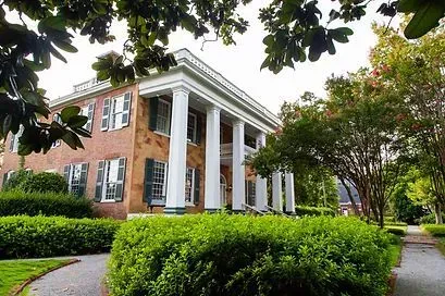 Brick building with white columns and large windows, surrounded by green shrubs and trees.