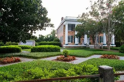 Elegant white-columned house with manicured green hedges, flower beds, and a brick walkway under trees