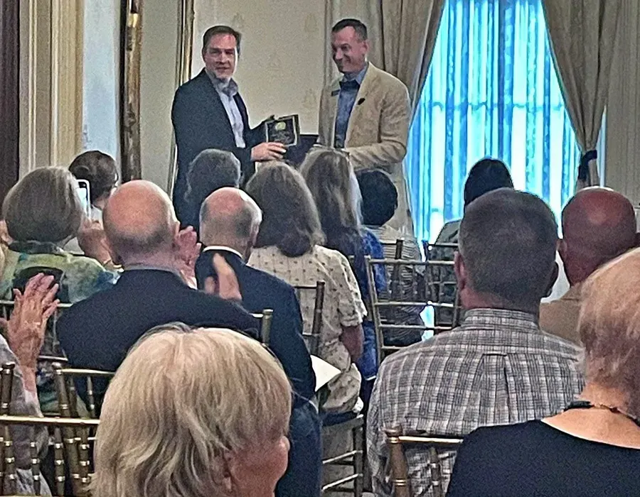 Two men at the front of a banquet room receive an item while seated guests applaud.