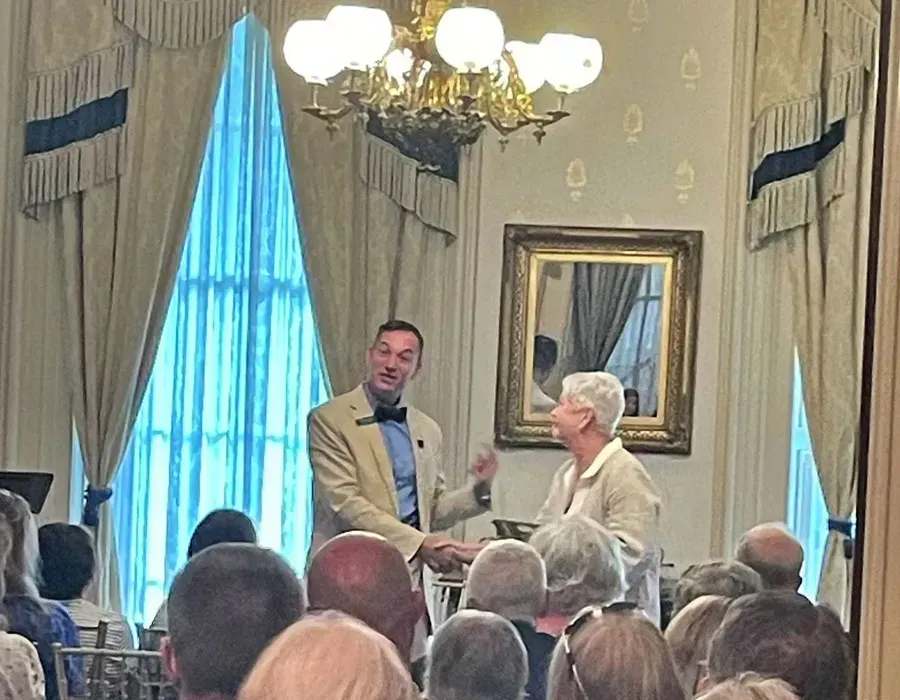 Man speaking at a podium to an audience in a formal room with chandeliers and tall drapes
