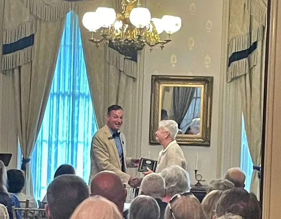 Man presenting a plaque to an elderly woman in a formal room with seated audience and chandeliers