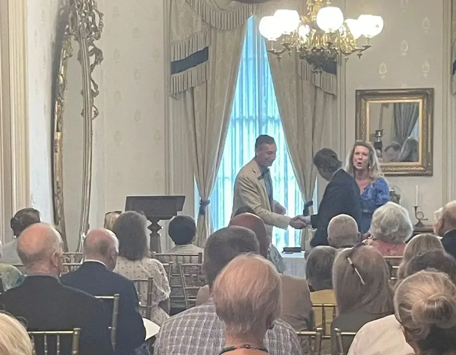 Audience seated in a formal room, watching a speaker at a podium near a bright window.